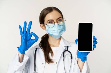 Online medical help concept. Enthusiastic young woman doctor in face mask, showing okay sign and mobile phone app, smartphone screen interface, white background