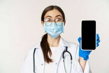 Online medical help concept. Surprised female doctor in face mask, looking amazed while showing mobile phone app, smartphone website, white background