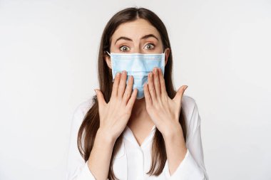 Covid and quarantine gesture. Young woman in face medical mask, looking surprised and shocked at camera, standing amazed against white background