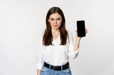 Şirket kıyafetleri içinde kendinden emin bir kadın, akıllı telefon ekranı gösteriyor, bir uygulamanın mobil arayüzü, beyaz arka planda duruyor.