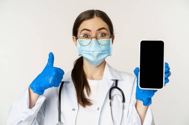 Online medical help concept. Enthusiastic young woman doctor in face mask, showing thumbs up and mobile phone app, smartphone screen interface, white background