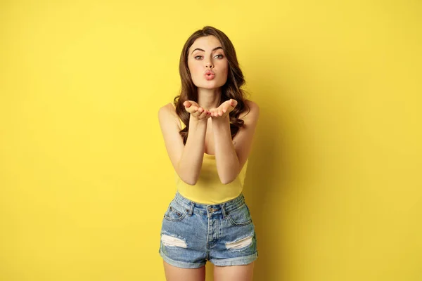 Beautiful, romantic brunette woman kissing, pucker lips, sending air kiss, mwah gesture, standing coquettish against yellow summer background — Zdjęcie stockowe