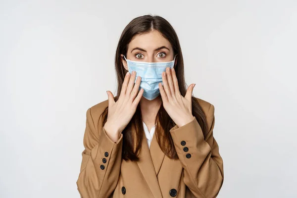 Workaplce and pandemic concept. Shocked business woman in face medical mask, gasping, looking startled and concerned at camera, standing over white background — Foto de Stock