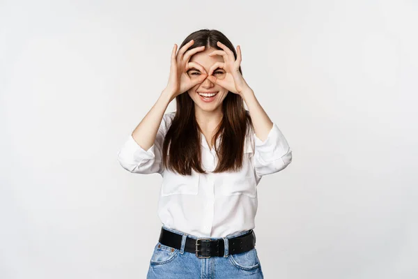 Funny girl looking through hand binoculars, zero gesture, standing over white background and smiling, seeing smth interesting far away, white background —  Fotos de Stock
