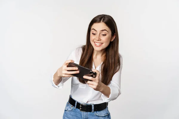 Mulher morena feliz jogando videogame móvel, sorrindo e olhando para a tela animada, em pé sobre o fundo branco — Fotografia de Stock