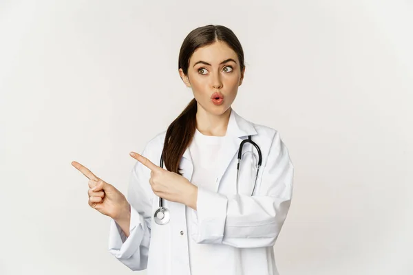 Surprised doctor, physician pointing fingers left and looking at logo banner, showing clinic advertisement, standing over white background — Stok fotoğraf