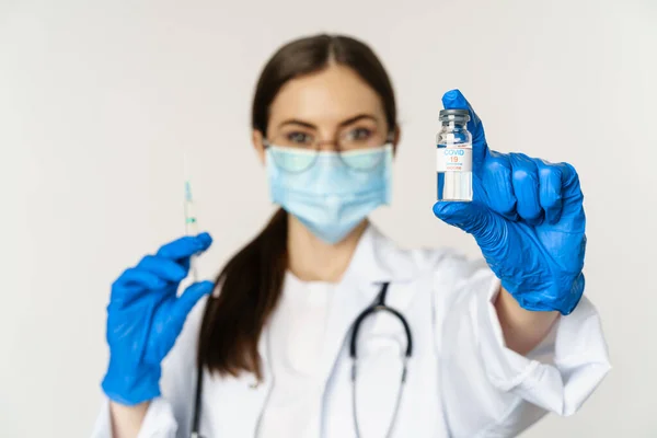 Covid-19 백신 과 건강 관리 개념. 의료용 마스크를 쓰고 있는 젊은 의사가 하얀 배경 위에 서 있는 코로나 바이러스 변형 백신 과 주사기를 보여 주고 있다 — 스톡 사진