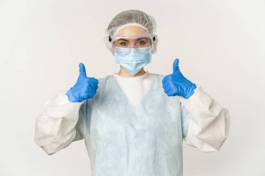 Şahsi koruyucu ekipman içinde doktor portresi, baş parmakları gösteren, yüz maskesi takan, covid-19 konsepti ve pandemik, beyaz arka plan