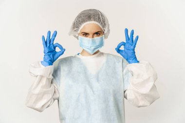 Image of healthcare worker, doctor in personal protective equipment from covid-19, showing okay ok sign, confident face in mask, standing over white background