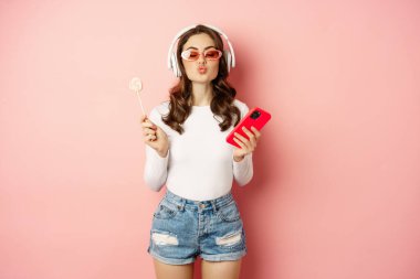 Stylish brunette woman dancing in headphones with mobile phone, listening music in earphones, licking lolipop, standing over pink background