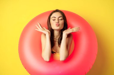Beautiful coquettish woman inside pink swimming ring, posing on summer vacation, beach holiday concept, standing against yellow background