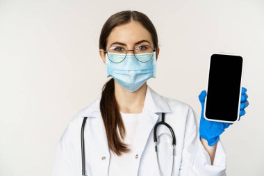 Çevrimiçi tıbbi yardım konsepti. Gözlüklü ve yüz maskeli kadın doktor, cep telefonu ekranını gösteriyor, hastalar için uygulama arayüzü veya web sitesi, beyaz arka planda duruyor