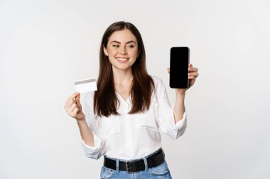 Portrait of female model showing credit card with smartphone screen, recommending application, standing over white background