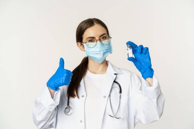 Profesyonel doktor, tıbbi maske ve gözlüklü doktor, aşıyı gösteren ve onaylayan, covid-19 'dan aşı olmayı öneren genç kadın portresi.
