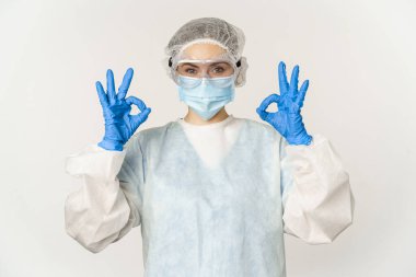 Doctor in personal protective equipment and face mask, showing okay, ok gesture, standing over white background