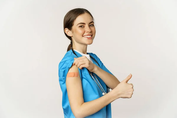 笑顔の看護師、親指と肩を示す医療従事者ワクチン接種後、 covid-19ワクチン接種キャンペーン、スクラブの白い背景の上に立って — ストック写真