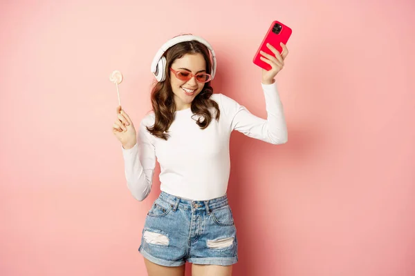 Dancing woman in sunglasses, listening music in headphones, holding lolipop and smartphone, laughing and smiling happy, standing over pink background — Zdjęcie stockowe