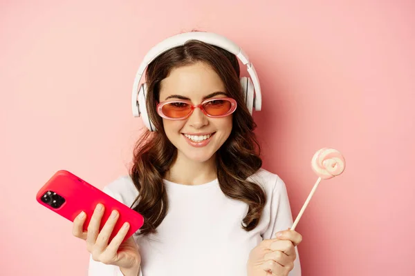 Close up portrait of stylish glamour girl, beautiful woman in headphones, listening music with smartphone, wearing sunglasses and licking lolipop — Zdjęcie stockowe