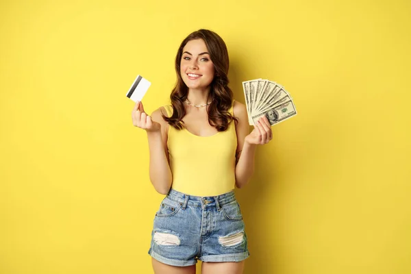 Jonge mooie vrouw in de zomer outfit met credit card en geld, contant geld in handen, staande tegen een gele achtergrond — Stockfoto