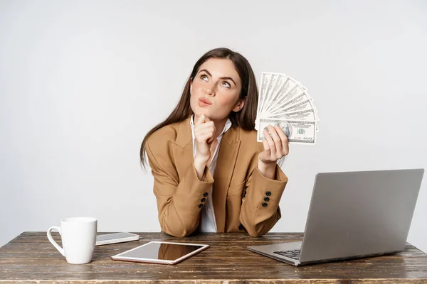Portrait of businesswoman sitting in office with money, working and making profit income, posing happy against white studio background — Stok fotoğraf