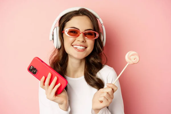 Stylish young woman with lolipop and cellphone, wearing sunglasses and headphones, listening music, standing over pink background — Zdjęcie stockowe