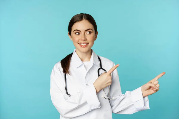 Porträt einer jungen Ärztin, einer Krankenhausmitarbeiterin, die mit dem Finger nach rechts zeigt, Logo zeigt, Klinik-Promo-Deal, vor torquosiertem Hintergrund steht — Stockfoto