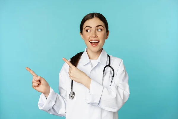 Retrato del trabajador médico sonriente, doctora en bata blanca con estetoscopio, señalando con los dedos a la izquierda, mostrando publicidad clínica médica, fondo turquesa —  Fotos de Stock
