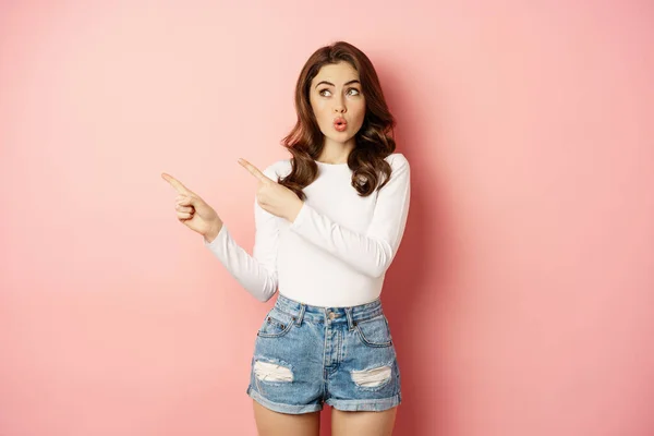 Wow, look there. Amazed feminine woman pointing fingers left, looking surprised and impressed, standing over pink background — Stock Photo, Image