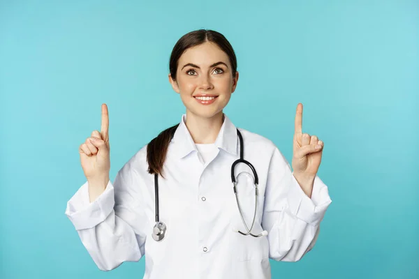 Enthusiastic medical worker, young woman doctor in white coat, stethoscope, showing advertisement, pointing fingers up, standing over torquoise background — Stock Fotó
