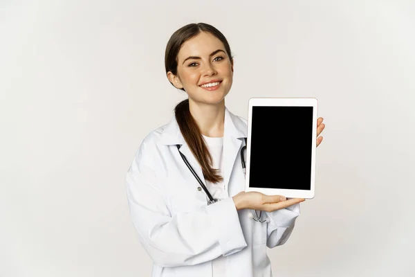 Ayuda médica en línea, cita remota. Sonriente hermosa mujer médico mostrando la pantalla de la tableta digital, demostrando el logotipo del sitio web, de pie sobre el fondo blanco — Foto de Stock