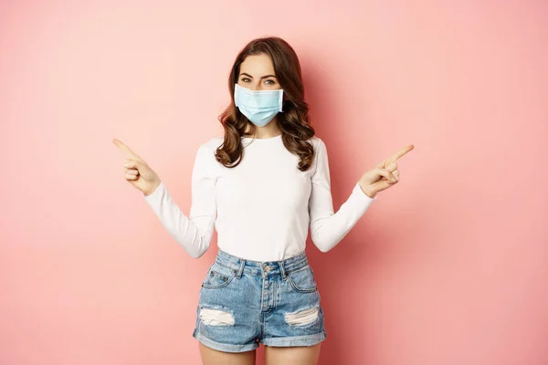 Portrait of beautiful woman in face medical mask pointing sideways, showing two choices, ways, sides, demonstrating advertisement, pink background — Fotografia de Stock