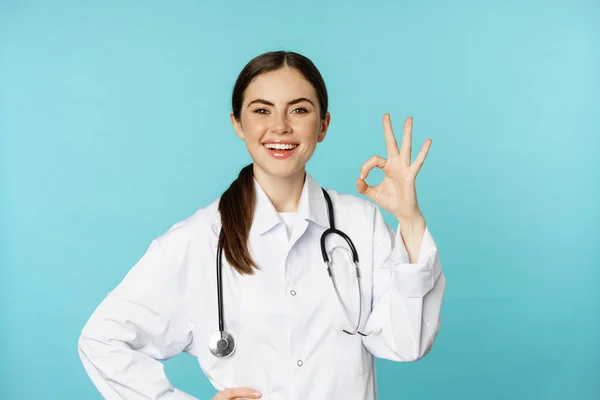 Porträt eines zufriedenen, lächelnden Arzthelfers, Ärztin zeigt Okay, ok, null Geste ohne Probleme, hervorragendes Zeichen, steht zufrieden über torquosiertem Hintergrund — Stockfoto