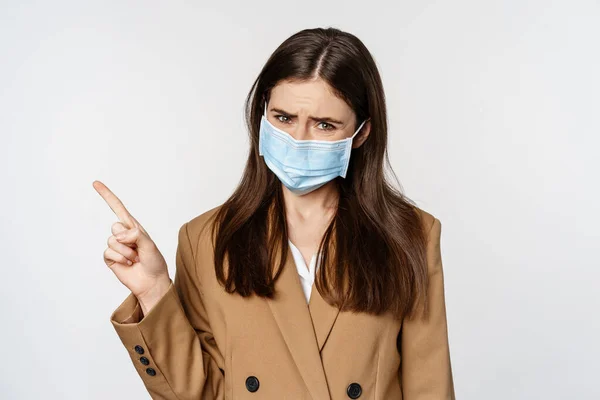 コロナウイルスと人々の概念。悲しいと懸念女性でオフィスの服や医療面マスク、動揺、左上隅に指を指す、白い背景 — ストック写真