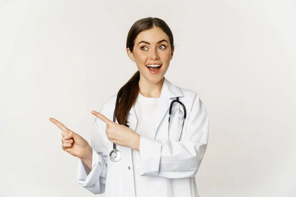 Surprised doctor, physician pointing fingers left and looking at logo banner, showing clinic advertisement, standing over white background — ストック写真