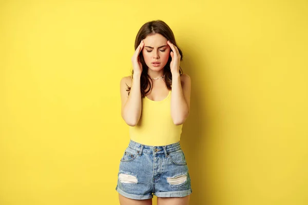 Stylish modern woman rubbing temples, having headache, migraine, frowning and grimacing from pain in head, standing against yellow background — Foto de Stock