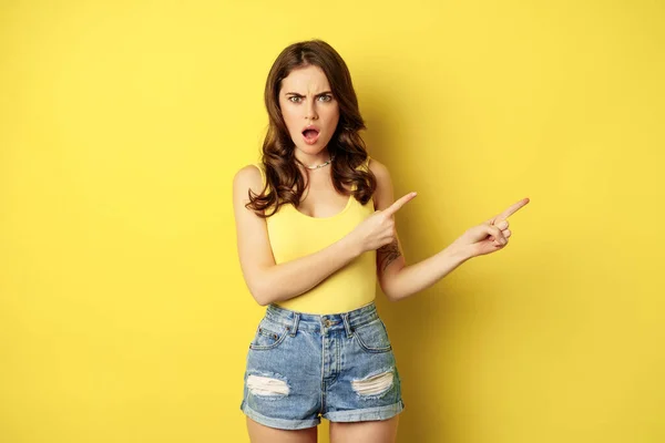 Shocked and frustrated young woman pointing right, showing banner or logo with angry, confused face expression, standing over yellow background — Foto Stock