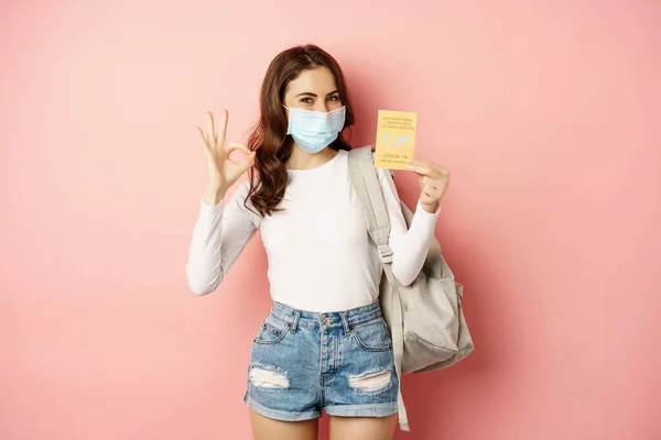 Νεαρή γυναίκα με ιατρική μάσκα, που δείχνει εντάξει σημάδι και covid πιστοποιητικό εμβολιασμού, ταξιδεύουν κατά τη διάρκεια πανδημίας, πρόκειται για διακοπές, στέκεται πάνω από ροζ φόντο — Φωτογραφία Αρχείου