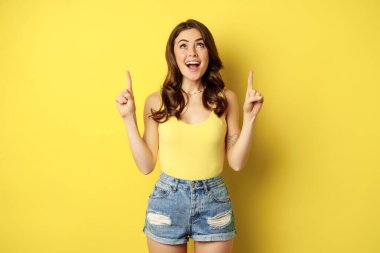 Portrait of cheerful girl smiling, pointing fingers up, showing promo text, summer advertisement, standing against yellow background