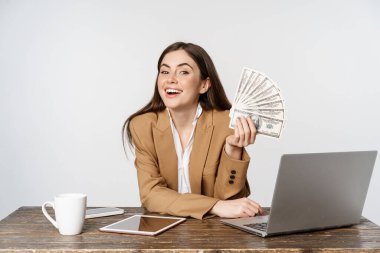 Portrait of businesswoman sitting in office with money, working and making profit income, posing happy against white studio background