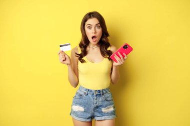 Stylish young brunette woman, holding credit card and smartphone, paying, purchase or order something in store, standing over yellow background