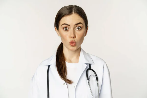 Retrato de médico mulher olhando surpreso, surpreso, reação de interesse e diversão, de pé no uniforme do hospital sobre fundo branco — Fotografia de Stock