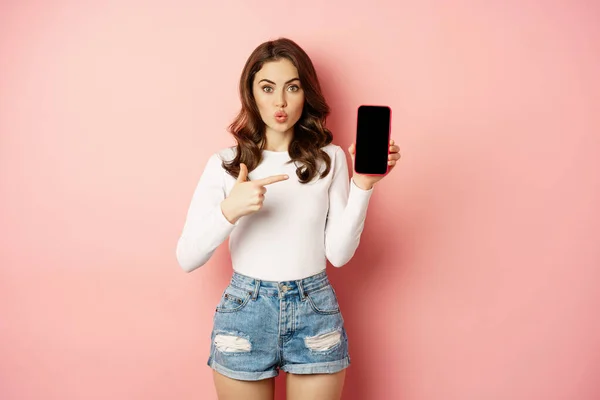 Zaskoczona brunetka wskazująca na ekran aplikacji telefonu komórkowego, pokazująca reklamę smartfona, ofertę zakupów online, stojąca zdumiona na różowym tle — Zdjęcie stockowe