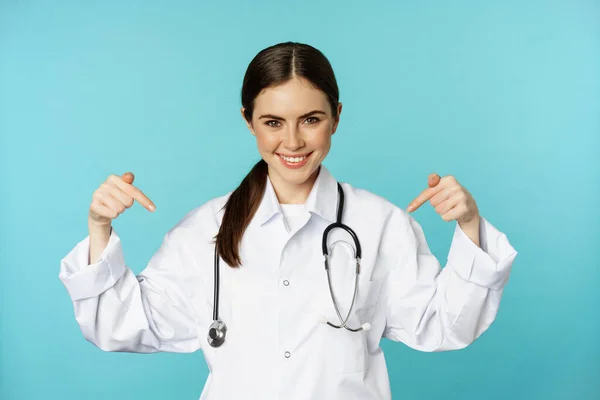 Smiling doctor medical worker, pointing fingers at logo, clinic banner, showing advertisement, wearing white coat, torquoise background — ストック写真