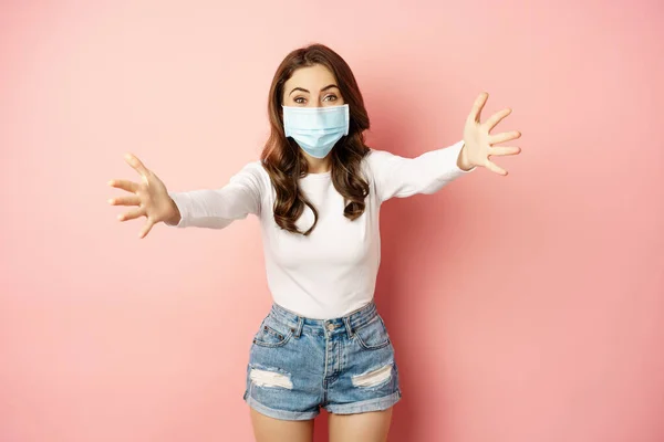 Portrait of smiling friendly girl in medical face mask, reaching hands, stretching arms forward for hug, receiving smth, standing over pink background — Φωτογραφία Αρχείου