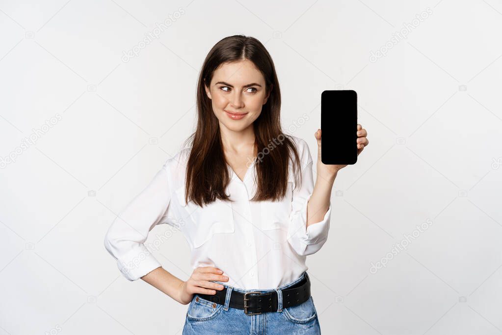 Smiling woman showing mobile phone screen, looking cunning, standing over white background