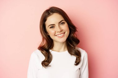 Close up of enthusiastic brunette girl with makeup, smiling and looking happy at camera, posing against pink background