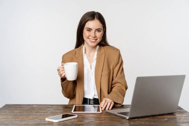Beautiful happy office lady, business woman drinking coffee at work, sitting at table and smiling pleased, holding white mug, studio background