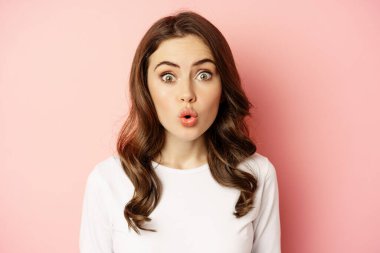 Image of stylish glamour woman looking surprised and amazed, saying wow with impressed reaction, standing over pink background
