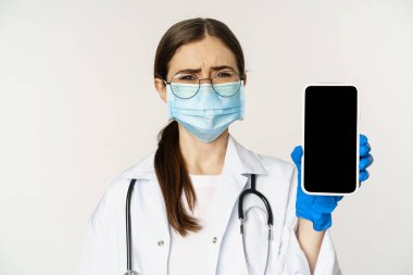 Online medical help concept. Sad and gloomy young doctor in face mask, showing smartphone screen with upset an disappointed expression, white background