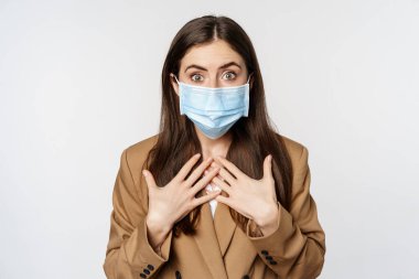 Workaplce and pandemic concept. Shocked business woman in face medical mask, gasping, looking startled and concerned at camera, standing over white background